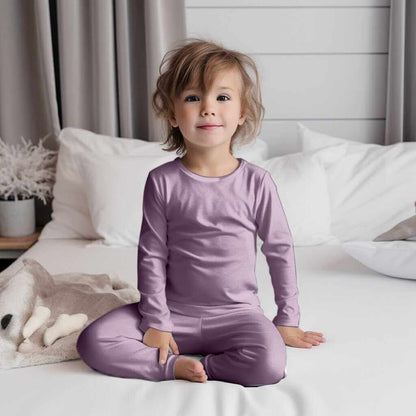 Smiling toddler on a bed wearing Wren Ivy Co. A Little Bit of Lavender in my Life bamboo pajamas, representing the brand's focus on sustainable and gentle baby wear.