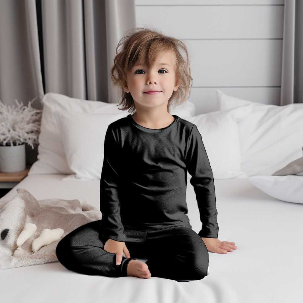 Cheerful toddler in Wren Ivy Co. black bamboo pajamas sitting cross-legged on a white bed, highlighting the softness and comfort of eco-friendly baby sleepwear.