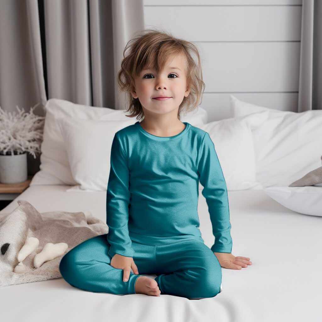 Happy toddler in Wren Ivy Co. teal bamboo pajamas sitting serenely on a bed, emphasizing the brand's commitment to sustainable baby clothing.