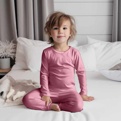 Smiling toddler in Wren Ivy Co. pink bamboo pajamas sitting on a white bed, accentuating the cozy and hypoallergenic qualities of the fabric.