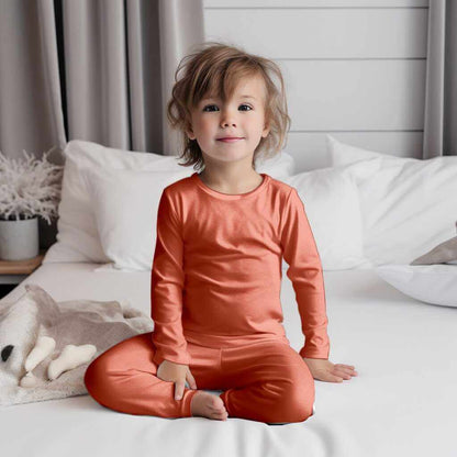 Toddler with a joyful expression wearing Wren Ivy Co. orange bamboo pajamas, sitting comfortably on a white bed, perfect for eco-friendly sleepwear.