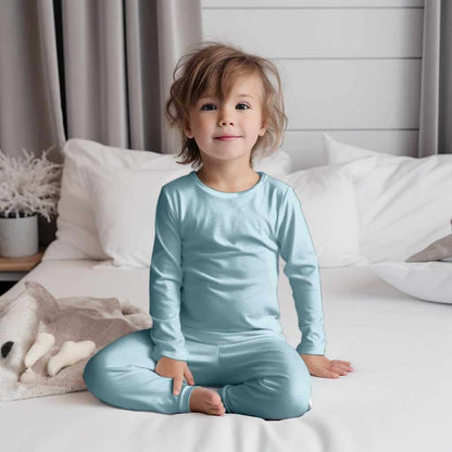 Cute toddler in Wren Ivy Co. baby Sad Beige Baby Blue bamboo pajamas sitting cross-legged on a bed, highlighting the brand's commitment to sustainable comfort.